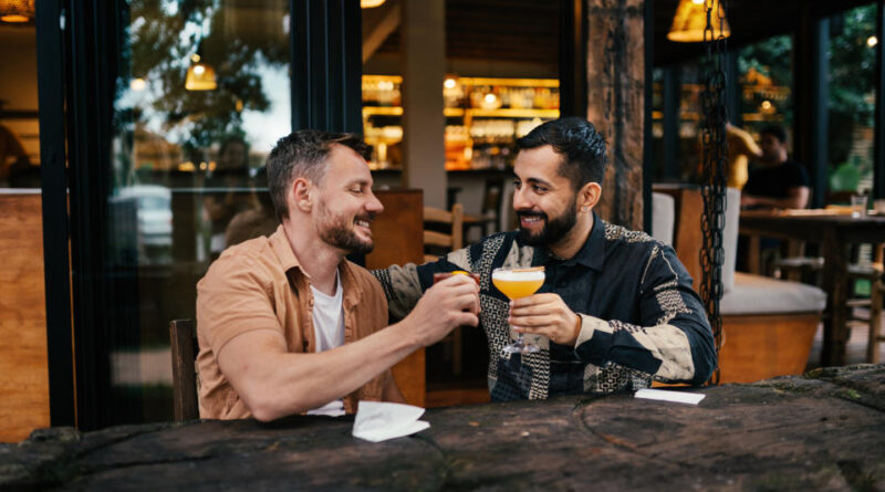 Young gay couple smiling while toasting cocktails together outside at a table in a bar square in the evening