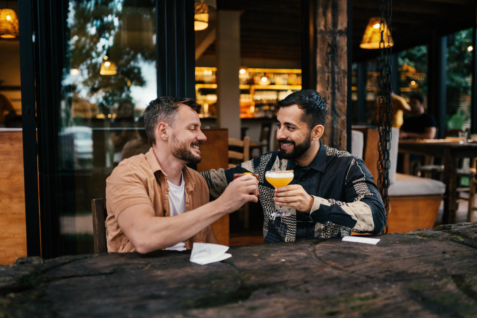 Young gay couple smiling while toasting cocktails together outside at a table in a bar square in the evening